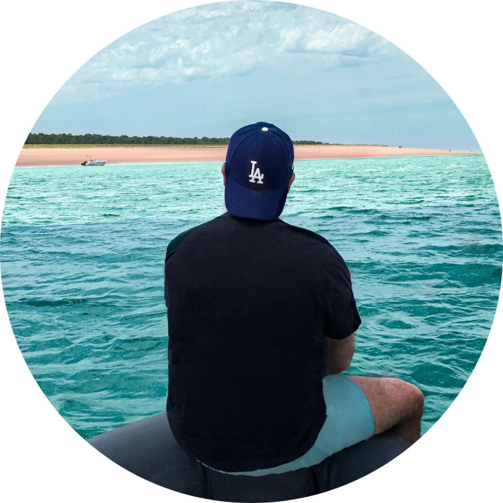 Image of man from behind sitting on a rock look at the water wearing a black shirt, and light blue pants with his navy cap backwards pondering about sperm donation