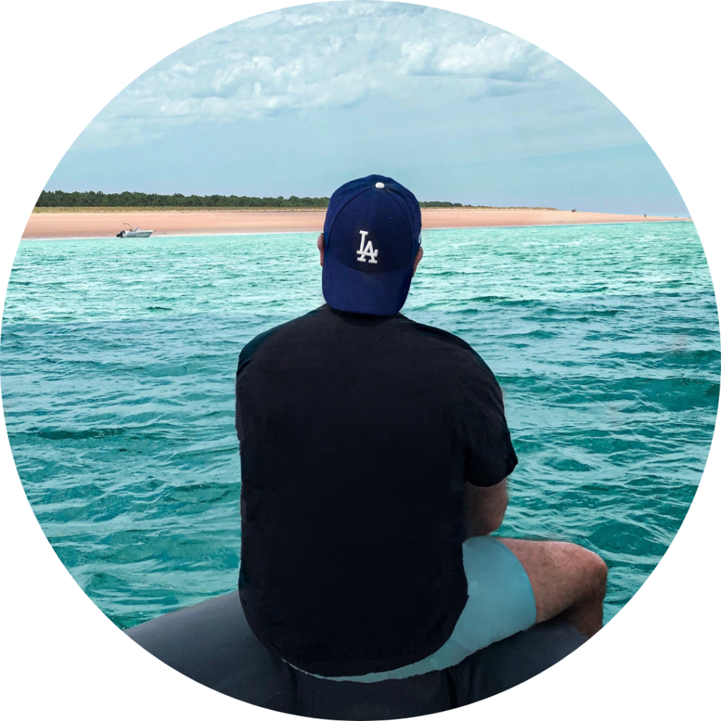 Image of man from behind sitting on a rock look at the water wearing a black shirt, and light blue pants with his navy cap backwards pondering about sperm donation